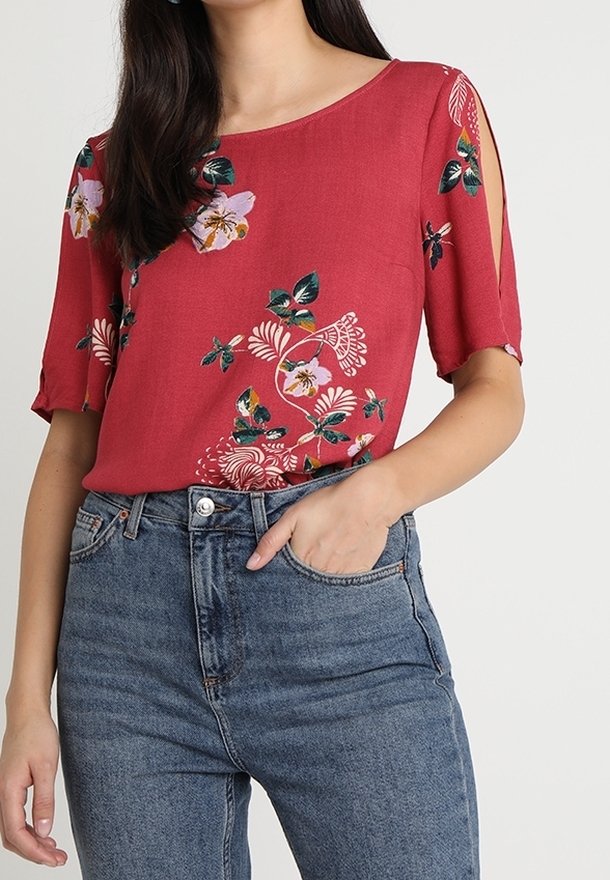 Femme portant un chemisier rouge à fleurs à manches courtes avec des découpes aux épaules et un jean bleu taille haute, main dans la poche avant.