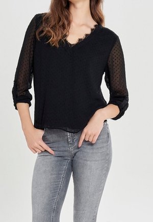 Woman wearing a black sheer polka-dot blouse with rolled-up sleeves and gray fitted jeans, posing with hands near pockets.