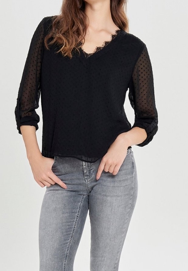 Femme portant un chemisier noir transparent à pois avec manches retroussées et un jean gris ajusté, posant avec les mains près des poches.