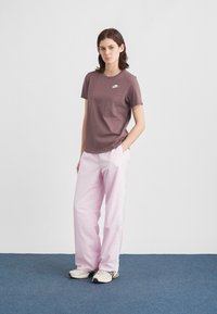 Νέα γυναίκα που στέκεται με τα χέρια στις τσέπες, φορώντας μωβ μπλουζάκι Nike, ανοιχτό ροζ παντελόνι και μπεζ αθλητικά παπούτσια πάνω σε μπλε χαλί.