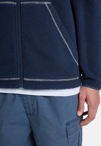 Μπλε ναυτική φλις ζακέτα με πλήρες φερμουάρ, υφή στην επιφάνεια και ραφές σε αντίθετο χρώμα. Φορεμένη πάνω από λευκό πουκάμισο και μπλε φορτηγατζή παντελόνια.