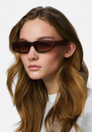 Fiatal nő hosszú hullámos hajjal, téglalap alakú teknősmintás napszemüveget és világosszürke felsőt visel, kissé oldalra nézve.