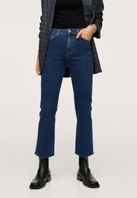 Mujer vestida con jeans azules de tiro alto y recortados con dobladillos deshilachados, botas negras hasta el tobillo, top negro y una blazer de cuadros, con una mano en el bolsillo.