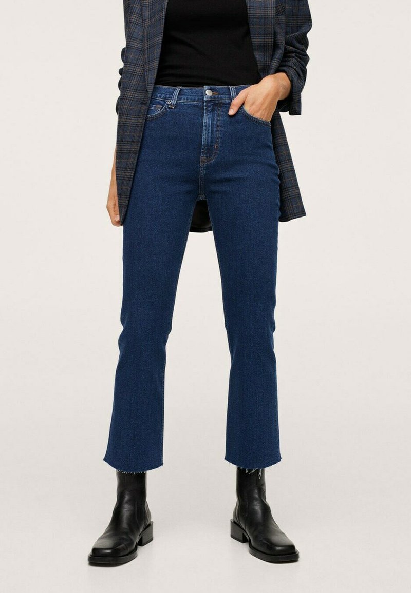 Mujer vestida con jeans azules de tiro alto y recortados con dobladillos deshilachados, botas negras hasta el tobillo, top negro y una blazer de cuadros, con una mano en el bolsillo.
