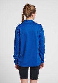 Maglia atletica a maniche lunghe di un blu vivace, realizzata in tessuto testurizzato. Presenta un colletto rialzato e una vestibilità leggermente ampia, abbinata a pantaloni corti neri.