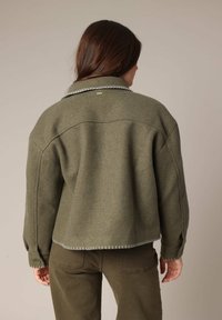 Veste en laine de couleur vert olive avec un design court, des coutures blanches le long de l'ourlet et un col pointu. Poignets boutonnés et accents minimaux.