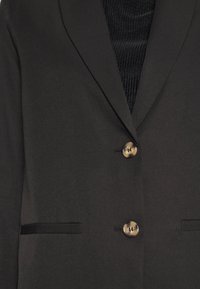 Blazer noir ajusté avec deux boutons beige et poches à rabats sur un top noir côtelé.