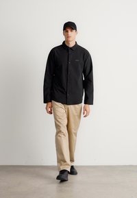 Ung mann som går, iført en svart skjorte med knapper, beige bukser, svart caps og svarte joggesko mot en ensfarget hvit bakgrunn.