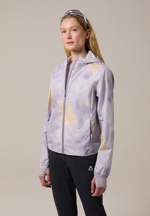 Mujer con chaqueta de teñido anudado de color lila claro con capucha, pantalones negros con logo y gafas de sol deportivas apoyadas en la cabeza, posando frente a un fondo liso.