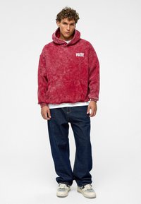 Rode tie-dye hoodie gemaakt van zachte stof, met een voorzak en witte "POLIZE" tekst. Combinant met donkerblauwe jeans en lichte sneakers.