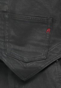 Bolso em denim preto com costura dupla e uma pequena letra "R" bordada em vermelho no lado direito, perto da borda superior.