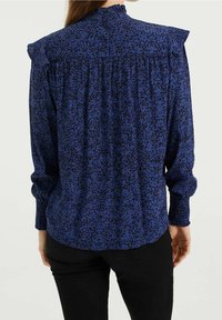 Žena v modré blůze s černým abstraktním vzorem, s volánovými rameny, řaseným zádovým dílem a dlouhými nařasenými manžetami, spárovaná s černými kalhotami.