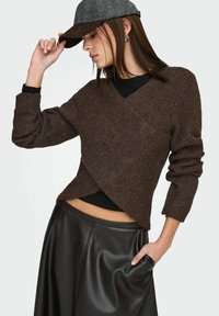 Pull marron tricoté avec col en V et design croisé, manches longues. Associé à une jupe en cuir noire, affichant une texture lisse.