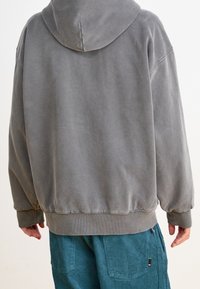 Grauer Kapuzenpullover mit lockerer Passform, gerippten Bündchen und Saum; der Stoff wirkt weich mit einer gebürsteten Textur. Getragen über teeblauem Cordhosen.