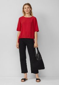 Haut rouge à manches courtes avec une couture froncée, associé à un pantalon noir cropped et un sac fourre-tout noir tissé, porté avec des sandales plates.