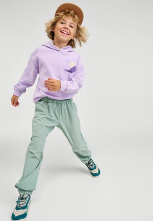 Uśmiechnięte dziecko z kręconymi włosami, ubrane w brązowy kapelusz, lawendową bluzę z kapturem, zielone spodnie i turkusowe trampki, idące do przodu na jednolitym tle.