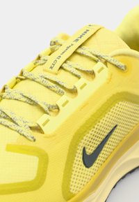 Zapatilla amarilla de Nike con tejido texturizado, paneles de malla y acentos grises. Presenta un sistema de cordones único y una marca en la lengüeta.