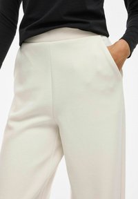 Světle béžové kalhoty vyrobené z hladkého materiálu, s elegantním designem, bočními kapsami a vysokým pasem, doplněné černým topem.