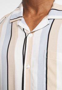 Camicia a maniche corte con righe verticali in beige, blu e bianco, dotata di bottoni frontali, tessuto morbido e taschino sul petto.