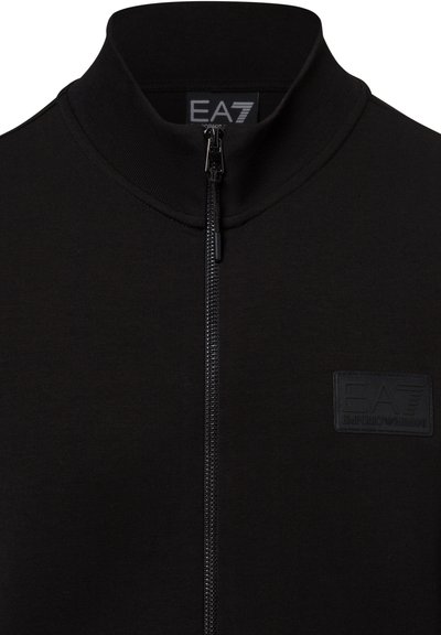 Schwarze Zip-Jacke mit hohem Kragen und EA7 Emporio Armani Logo-Patch auf der Brust sowie Etikett im Kragen.