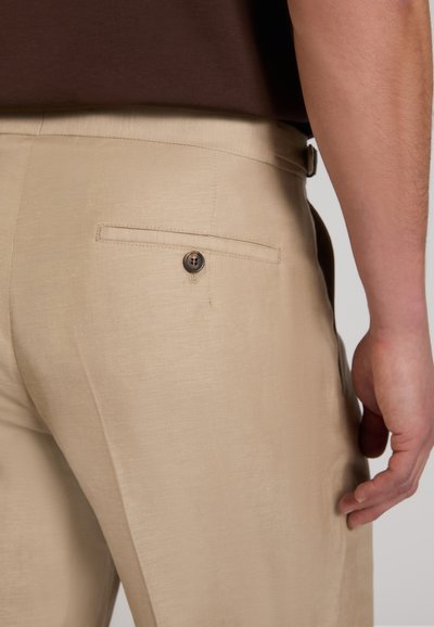 Close-up of beige pants with a buttoned back pocket worn by a person standing with their right arm relaxed at their side.