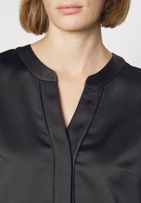 Femme aux cheveux courts brun clair portant un chemisier en satin noir avec un col rond et un pli central à l'avant.