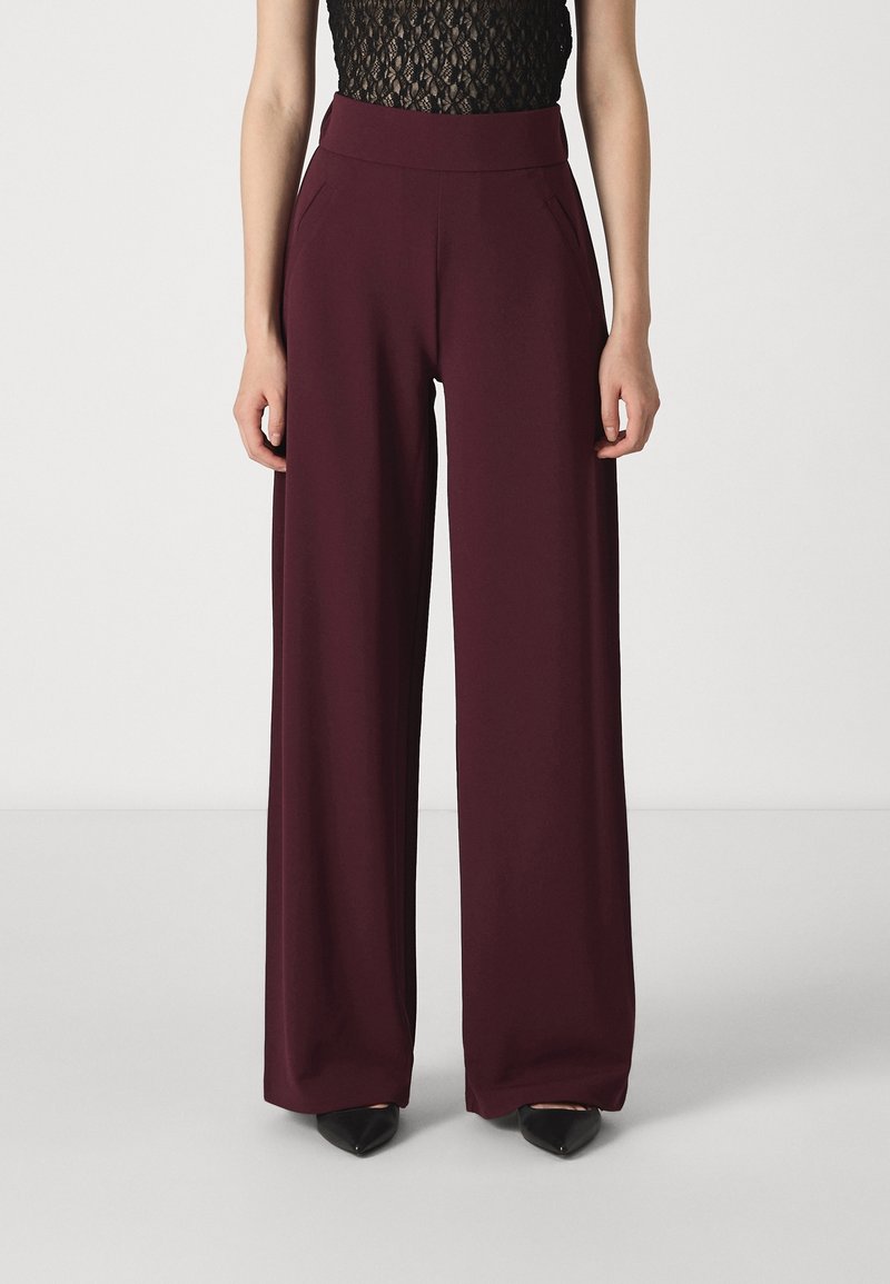 Femme portant un pantalon bordeaux taille haute à jambes larges et un haut sans manches en dentelle noire, debout devant un fond uni.