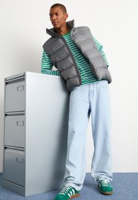 Grå puffervest over en grøn og hvid stribet skjorte, lyseblå vidde jeans og grønne sneakers med hvide striber, lænet op ad et gråt arkivskab.