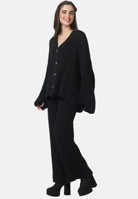 Zwarte, geribbelde gebreide cardigan met een V-hals, knoopsluiting en oversized mouwen, gecombineerd met bijpassende geribbelde wijde broek en plateauschoenen.