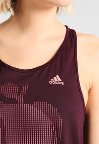 Débardeur athlétique bordeaux avec un motif à pois roses, un col rond et un petit logo Adidas rose en haut à gauche. Texture du tissu douce.