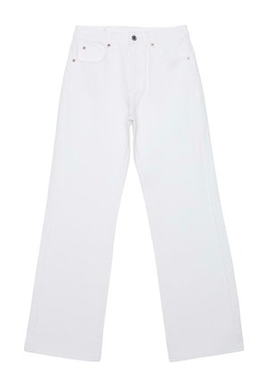 Jeune femme aux cheveux blonds bouclés porte un caraco en dentelle noire, un pantalon blanc à jambes larges, tient un sac noir à franges, debout sur fond gris.