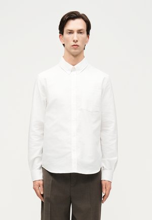 Άνδρας που στέκεται μπροστά σε ένα απλό φόντο φορώντας ένα λευκό πουκάμισο με κουμπιά και σκούρο καφέ παντελόνι, κοιτάζοντας προς τα εμπρός με ουδέτερη έκφραση.