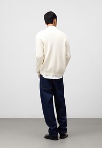Maglione a coste crema con colletto alto, abbinato a jeans in denim scuri dal fit largo e scarpe nere. Sfondo semplice.