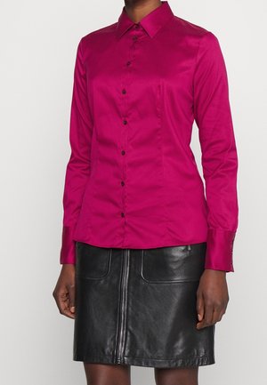 Person trägt ein eng anliegendes magentafarbenes Hemd mit Knöpfen und langen Ärmeln sowie einen schwarzen Lederminirock mit Reißverschluss vorne.