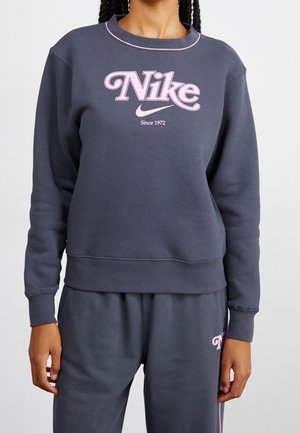 Person wearing dark gray Nike sweatshirt and sweatpants with pink logo and trim, standing against a plain light background.