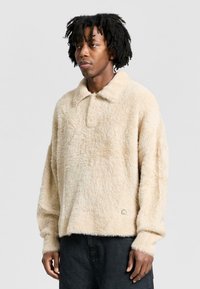 Pullover beige peloso con colletto, caratterizzato da una superficie testurizzata. Appropriato e ampio con maniche lunghe, presenta un piccolo logo in basso.