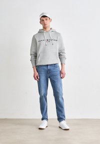 Gray hoodie with logo, drawstrings, and kangaroo pocket. Light blue jeans, white sneakers with light brown soles, and a white cap.
