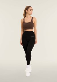 Top sportivo corto marrone con logo, abbinato a leggings neri a vita alta con logo. La modella indossa scarpe sportive bianche e calze.