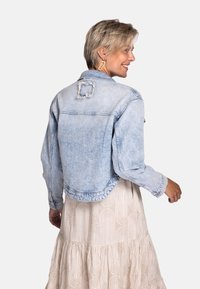 Hellblaue Jeansjacke mit langen Ärmeln, die auf der Rückseite einen verfaulten Fleck und einen abgerundeten Saum aufweist. Getragen über einem gemusterten, fließenden Rock.