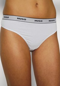 Sous-vêtements en coton blanc de style bikini avec une bande élastique grise portant le logo "HUGO" en noir. Texture douce et coupe ajustée.