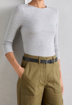 Person trägt ein hellgraues, geripptes Langarmshirt, das in eine hoch taillierte olivgrüne Hose mit schwarzem Gürtel gesteckt ist und die Hände in den Taschen hat.