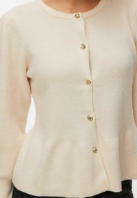 Cardigan en maille côtelée beige à manches longues, orné de cinq boutons dorés décoratifs en forme de fleurs sur le devant, porté avec un pantalon foncé.