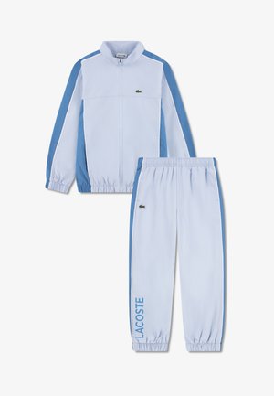Jachetă sport de culoare albastru deschis, cu accente albastre și închidere cu fermoar, asortată cu pantaloni potriviți. Dispune de manșete elastice și inscripția „LACOSTE” pe picior.