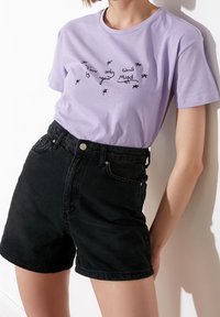 Camiseta de algodón lavanda con texto negro bordado, combinada con pantalones cortos de mezclilla negros de talle alto, que cuentan con bolsillos frontales y un cierre de botón.