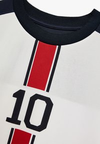 Valkoinen urheilujersey, jossa on tummansiniset ribbikaulus, punainen pystyraita ja numero "10" mustalla. Teksturoitu kangas.