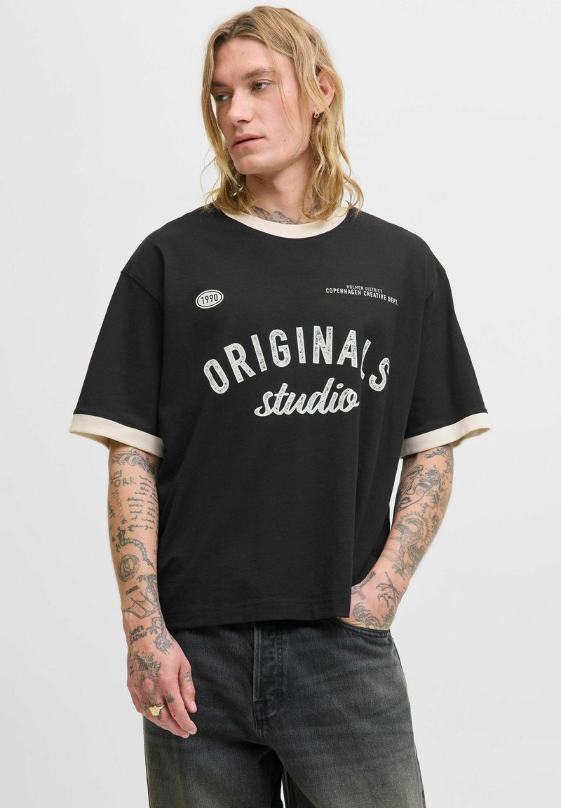 Camiseta negra de manga corta con ribete beige, que presenta "ORIGINALS studio" en letras gráficas blancas y un pequeño logo, hecha de algodón suave.