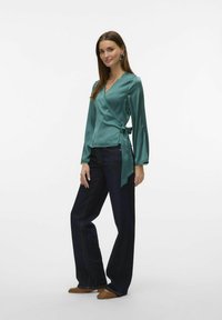 Teal satin indpakningsbluse med lange ærmer, bånddetalje i taljen, parret med mørkeblå vide bukser og brune flade sko.