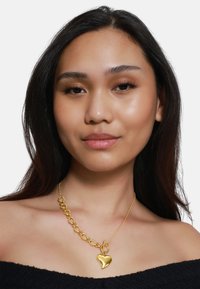 Collana con pendente a forma di cuore in oro, caratterizzata da un design con catena spessa. La catena presenta grandi anelli, mentre il pendente a forma di cuore ha una superficie liscia e riflettente.