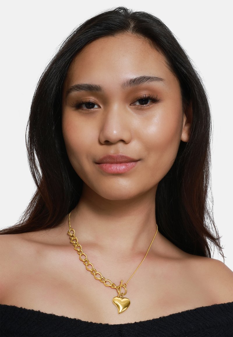 Collana con pendente a forma di cuore in oro, caratterizzata da un design con catena spessa. La catena presenta grandi anelli, mentre il pendente a forma di cuore ha una superficie liscia e riflettente.