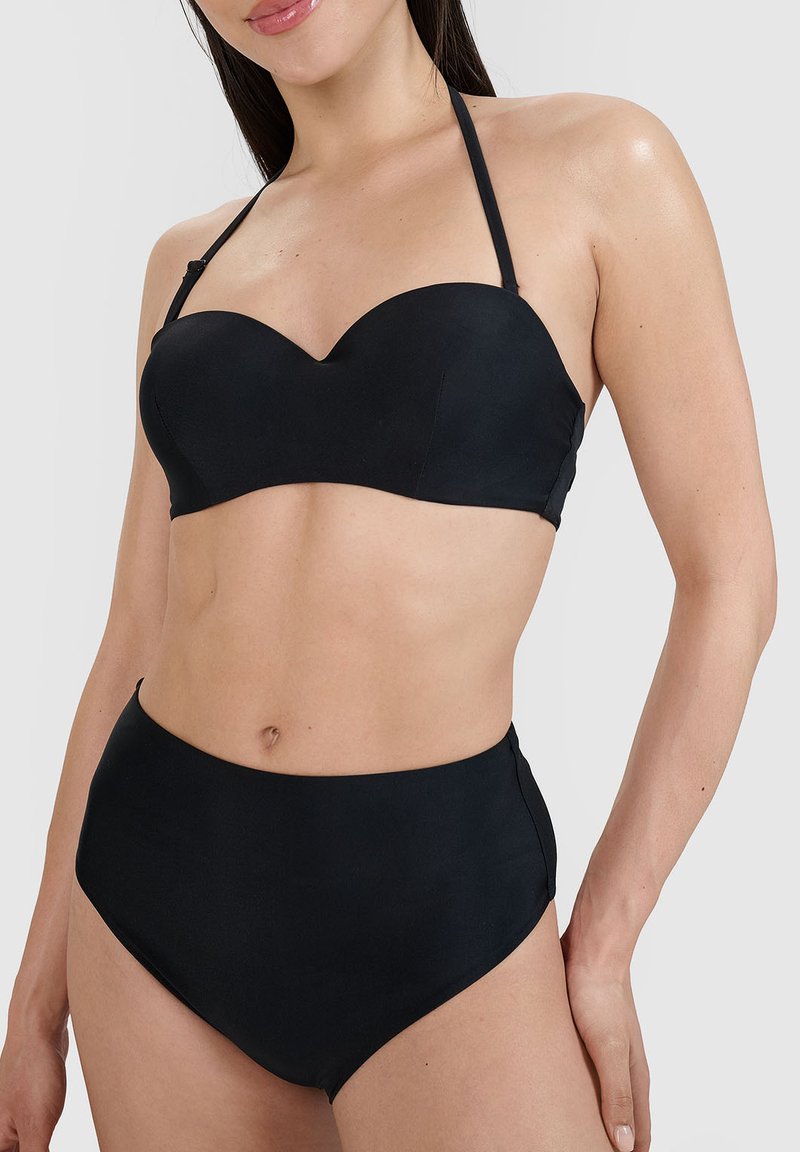 Femme portant un bikini noir taille haute avec un haut bustier sans bretelles en forme de cœur et une bride autour du cou sur un fond uni.